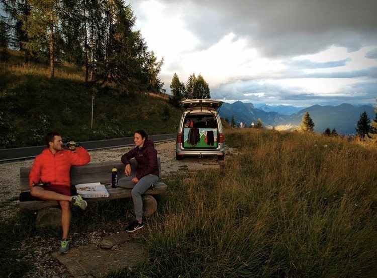 People eating pizza on bench in front of camper in mountains.