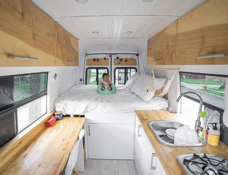 Custom van ideas - white interior of van with bed at back and wooden work top and table on either side.
