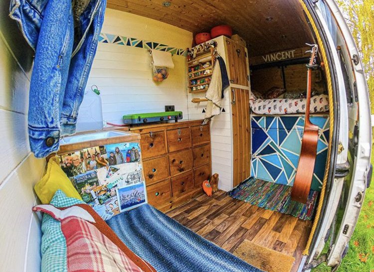 Van interior with wooden and white walls and funky pattern on drawers beneath fixed bed at back.